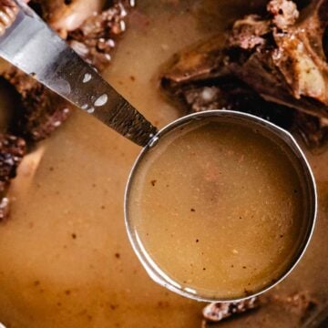 Ladle of warm beef bone broth lifted from a pot with roasted bones.
