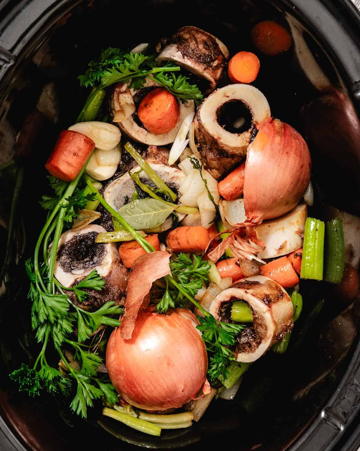 Slow cooker filled with roasted beef bones, carrots, celery, onions with skins, garlic, bay leaves, and fresh herbs before adding water for bone broth.