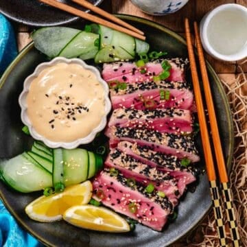 Seared ahi tuna with sesame dipping sauce with cucumber and lemon wedges. On a plate with chopsticks, with sake on the table.