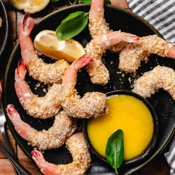 Baked coconut shrimp on a black plate with lemon wedges and a bowl of mango dipping sauce