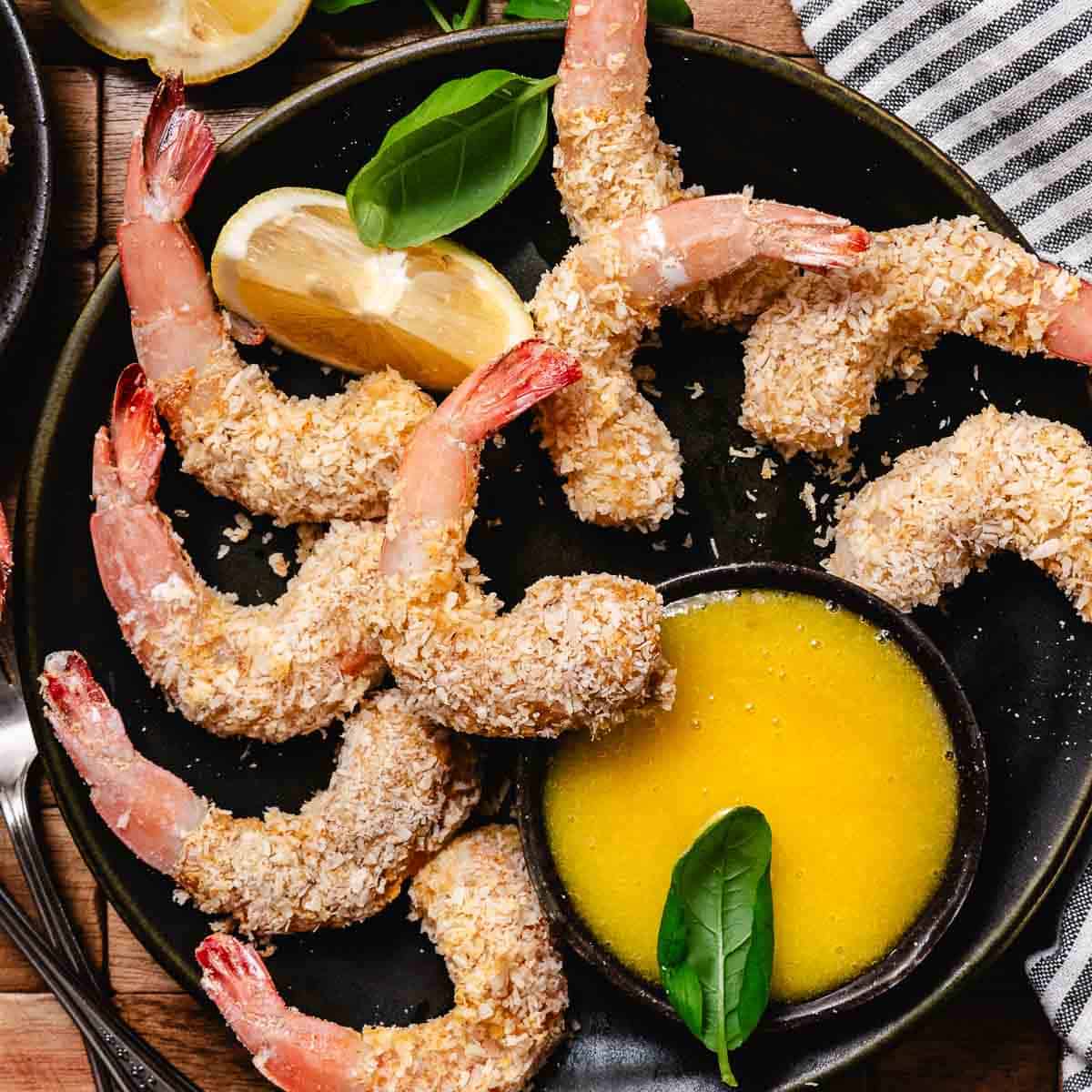 Baked coconut shrimp on a black plate with lemon wedges and a bowl of mango dipping sauce