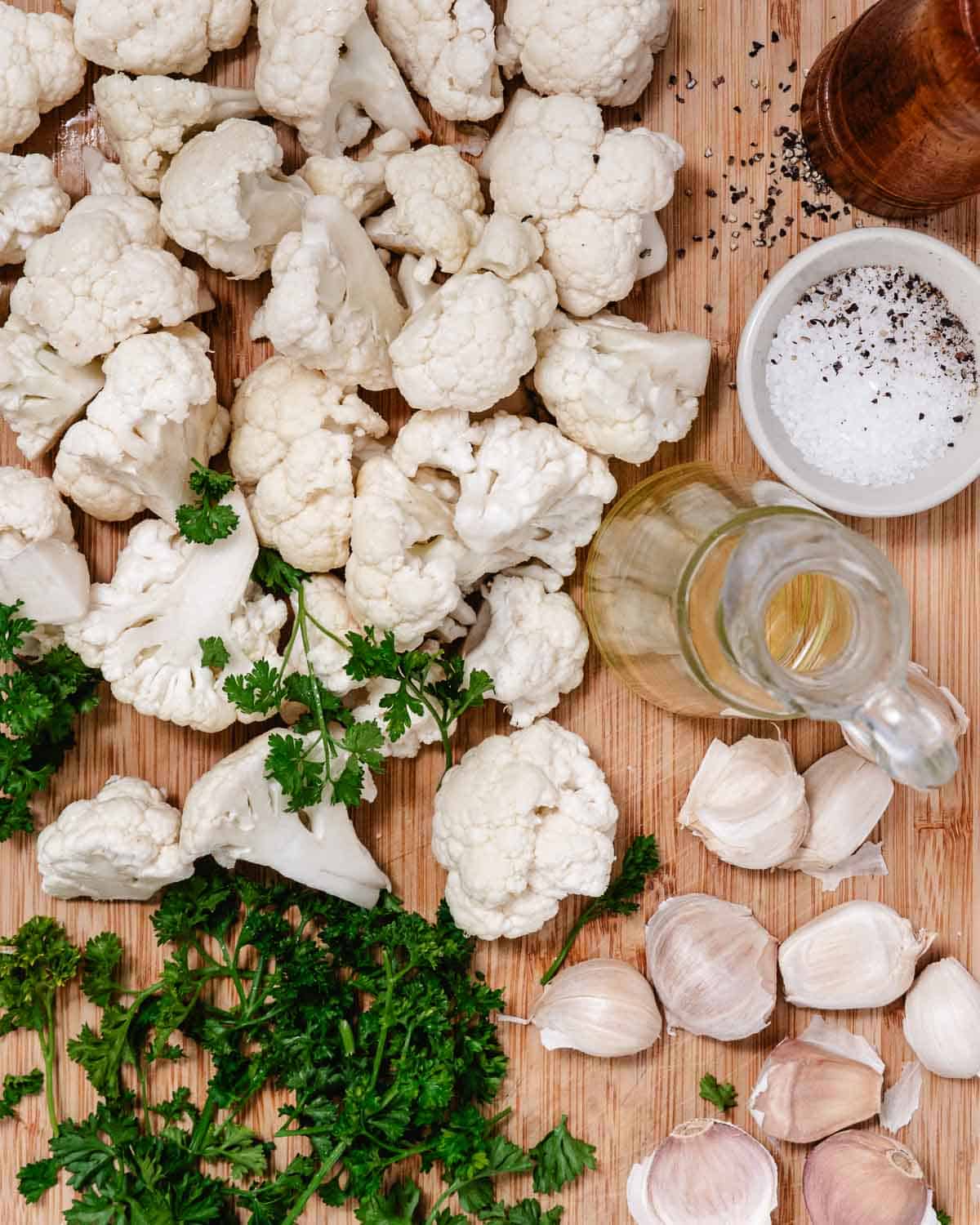 Raw cauliflower florets with whole garlic cloves, olive oil, kosher salt, black pepper, and fresh parsley arranged on a wooden cutting board.