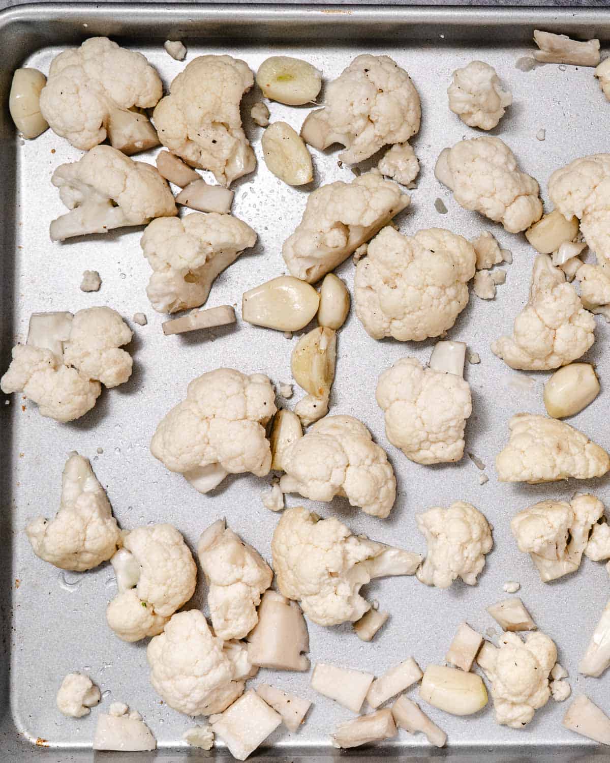 Cauliflower florets, chopped stems, and whole garlic cloves spread in a single layer on a metal sheet pan before roasting.