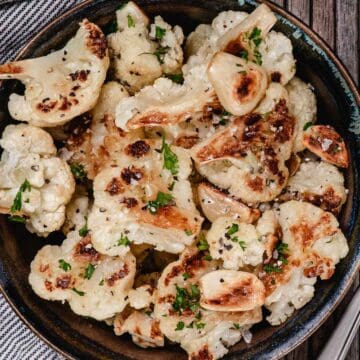 Oven-roasted garlic cauliflower with golden brown edges, whole caramelized garlic cloves, chopped parsley, salt, and black pepper.