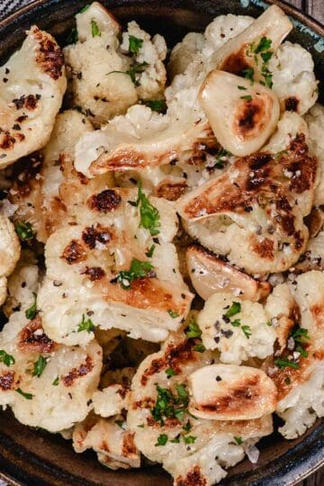 Oven-roasted garlic cauliflower with golden brown edges, whole caramelized garlic cloves, chopped parsley, salt, and black pepper.