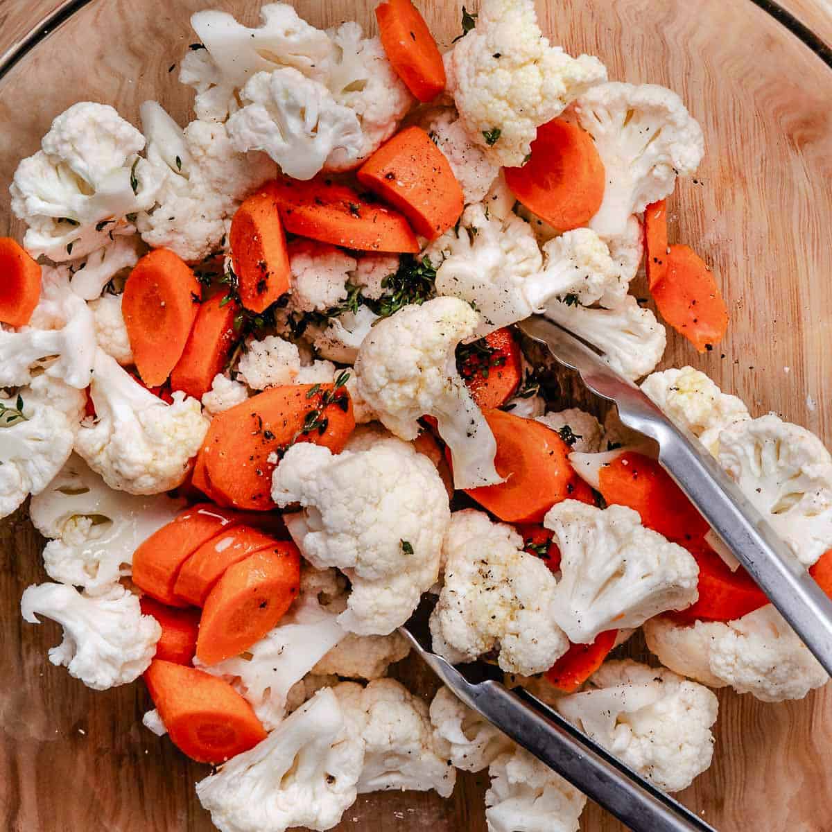 Mixing cauliflower, carrots, and thyme with tongs in a bowl before roasting,