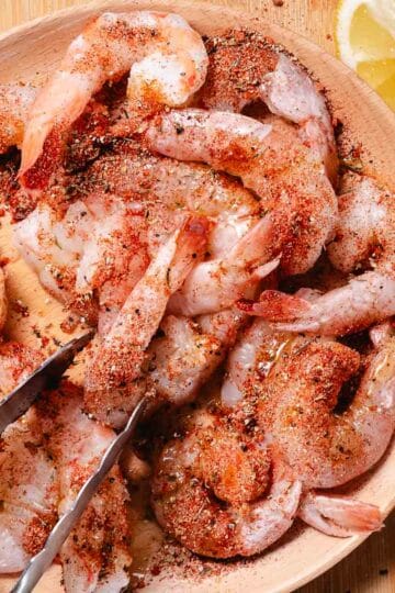 Raw shrimp tossed with blackened seasoning in a wooden bowl with lemon wedges on the side.