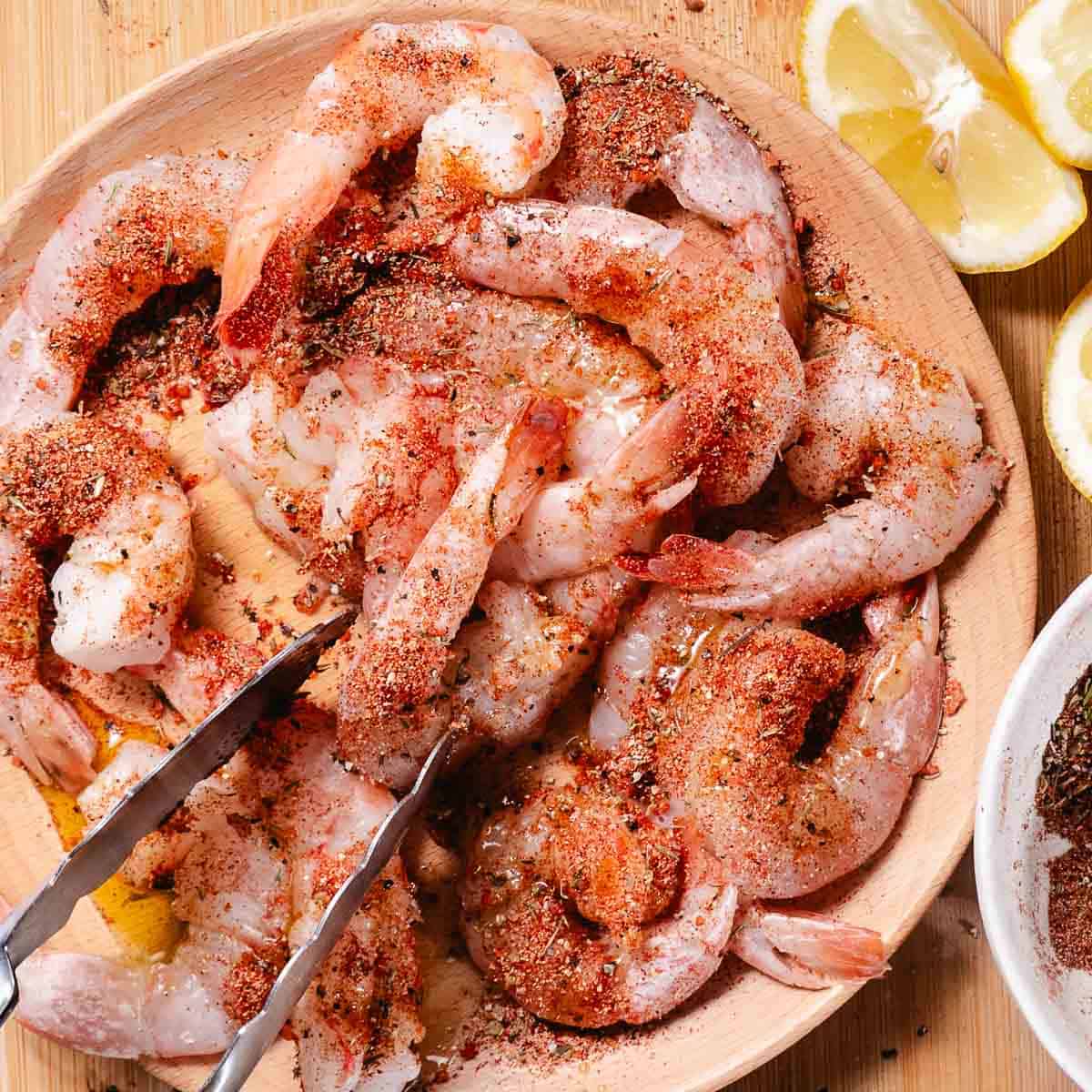 Raw shrimp tossed with blackened seasoning in a wooden bowl with lemon wedges on the side.