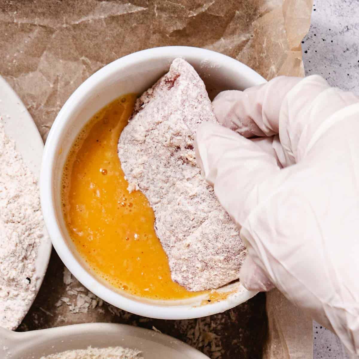 Floured red snapper fillet being dipped into beaten egg, the second step before coating in panko