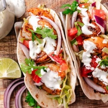 Shrimp tacos with seasoned shrimp, lettuce, tomatoes, red onion, cilantro, creamy taco sauce, and lime in warm tortillas.