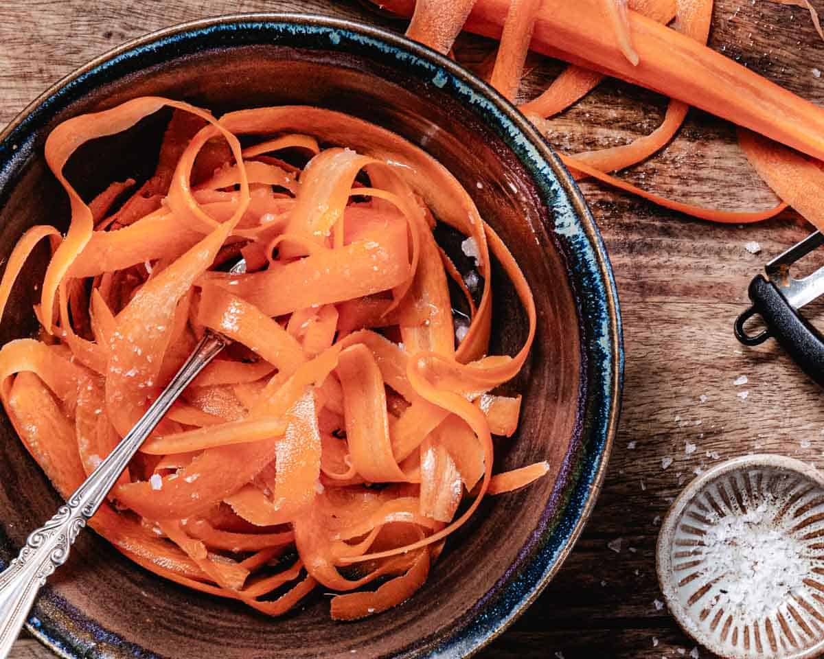 fresh carrot salad with flaky salt in a dark ceramic bowl on a rustic wood table
