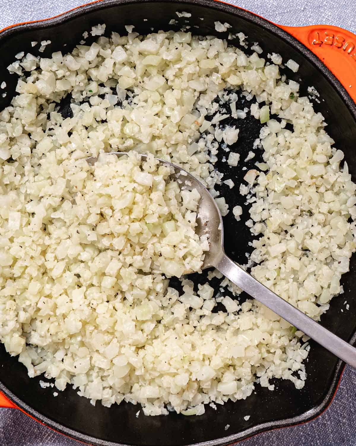 Finely chopped cauliflower in a skillet, ready to be cooked into cauliflower rice, on a cooking surface.