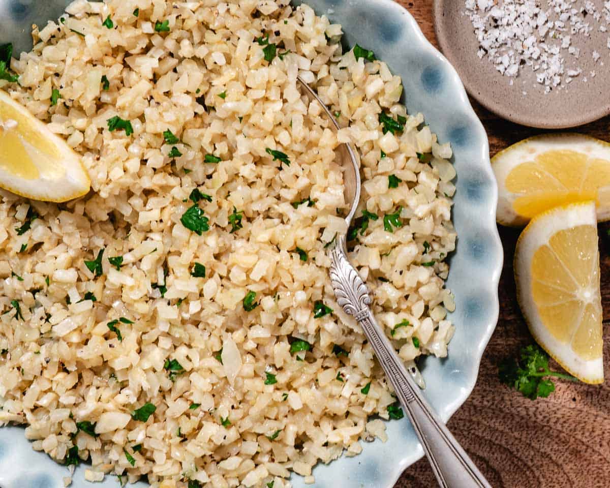 Lemon parsley cauliflower rice in a serving bowl with fresh lemon wedges.