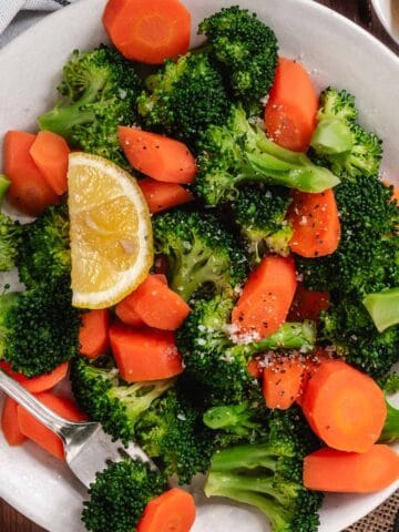 Steamed broccoli and fresh carrots on a white plate with a lemon slice.