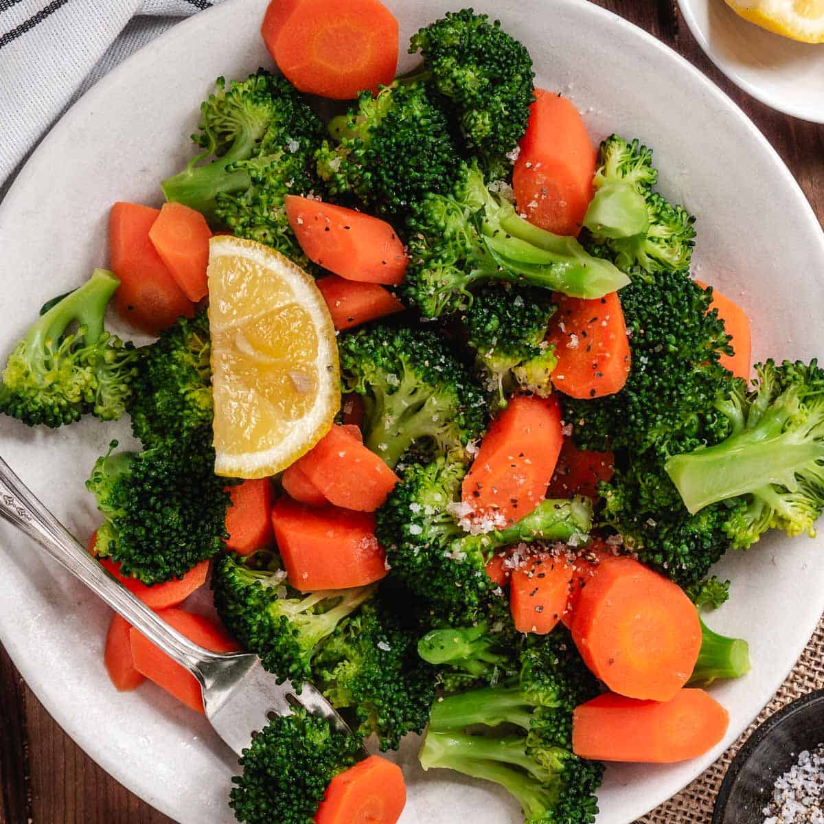 Steamed broccoli and fresh carrots on a white plate with a lemon slice.