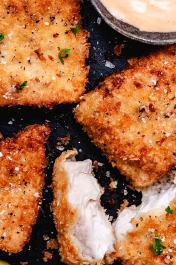 Crispy pan-fried mahi mahi fillets with panko breadcrumbs, flaky sea salt, parsley, lemon wedges, and creamy bang bang sauce on a dark slate background.