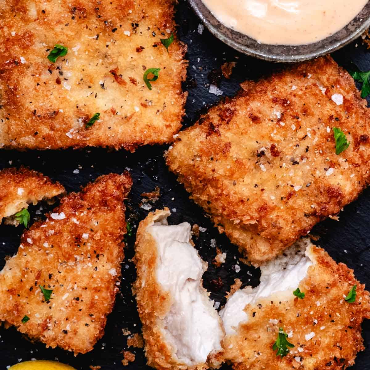 Crispy pan-fried mahi mahi fillets with panko breadcrumbs, flaky sea salt, parsley, lemon wedges, and creamy bang bang sauce on a dark slate background.