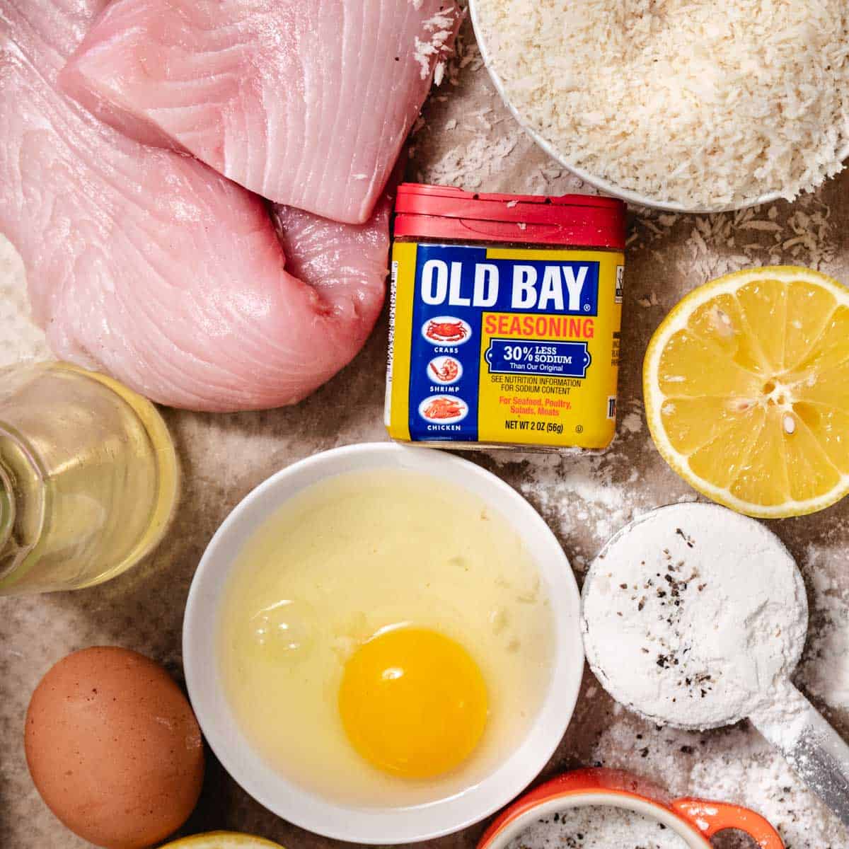 Ingredients for pan-fried mahi mahi including fresh mahi fillets, panko breadcrumbs, Old Bay seasoning, egg, flour, lemon, and vegetable oil arranged overhead.