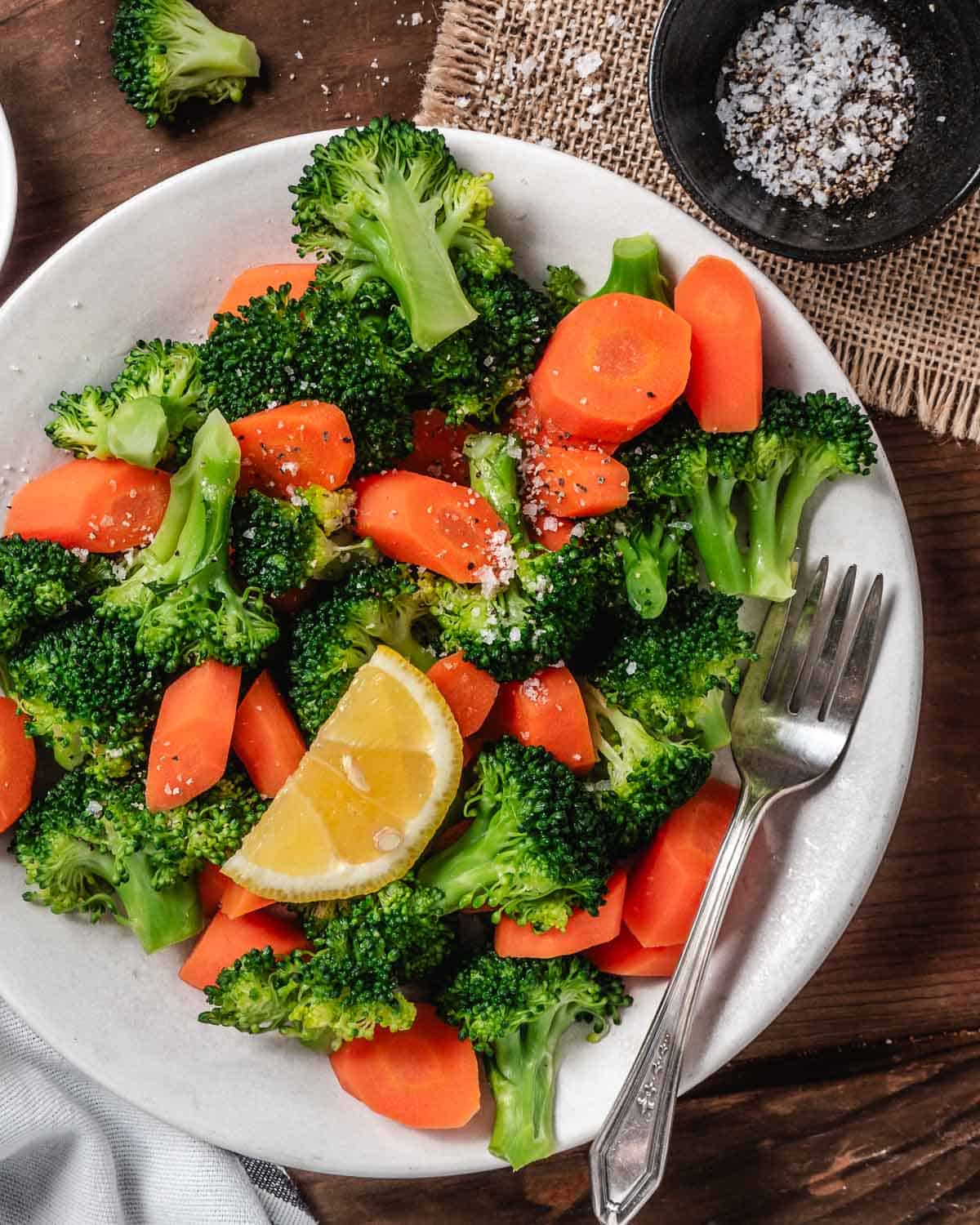 Cooked broccoli and carrots on a plate with a lemon wedge.
