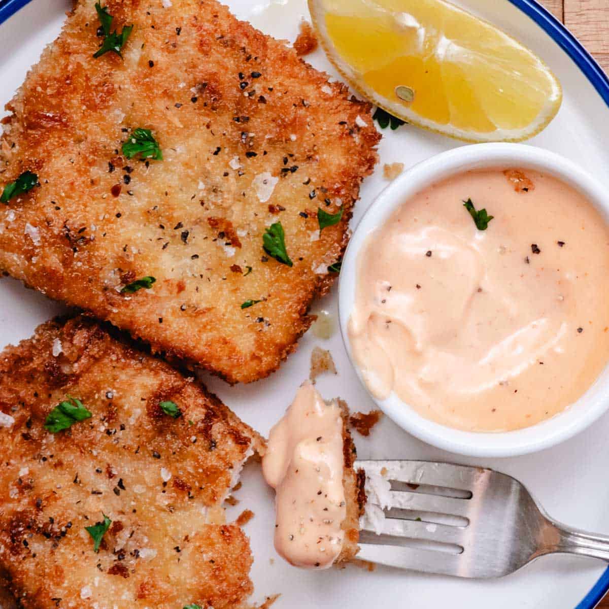 Fork holding a bite of crispy fried mahi mahi dipped in creamy bang bang sauce with lemon wedges on a white plate.