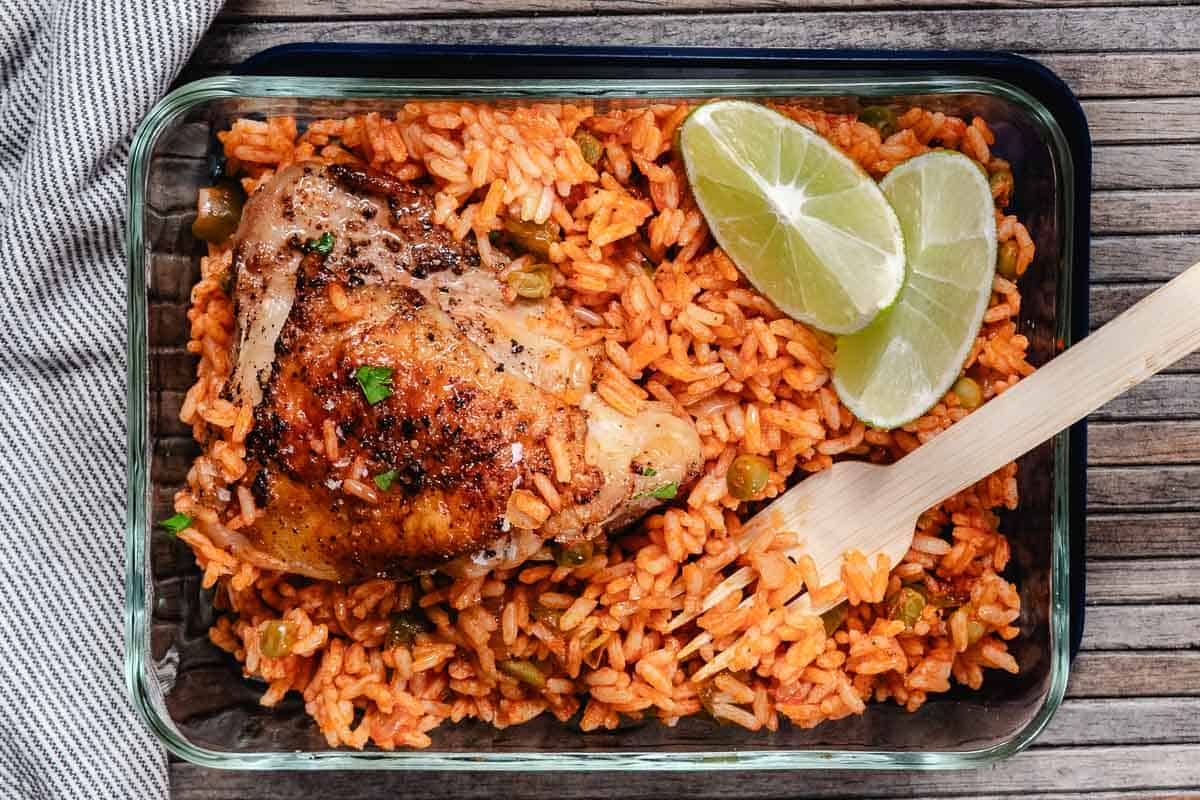 Spanish rice with peas topped with roasted chicken and lime wedges in a glass meal-prep container.
