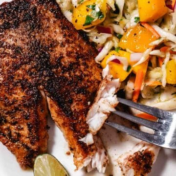 Fork pulling apart a flaky pan-seared mangrove snapper fillet served with mango cabbage slaw and lime.