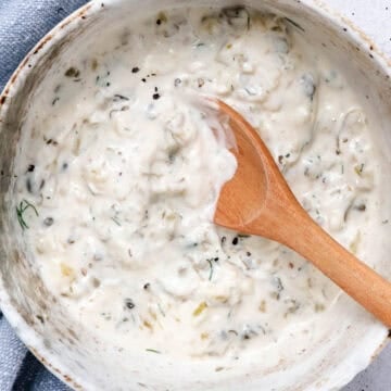 Homemade tartar sauce with dill, capers, pickles, and onion in a ceramic bowl with a wooden spoon.