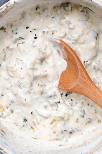 Homemade tartar sauce with dill, capers, pickles, and onion in a ceramic bowl with a wooden spoon.