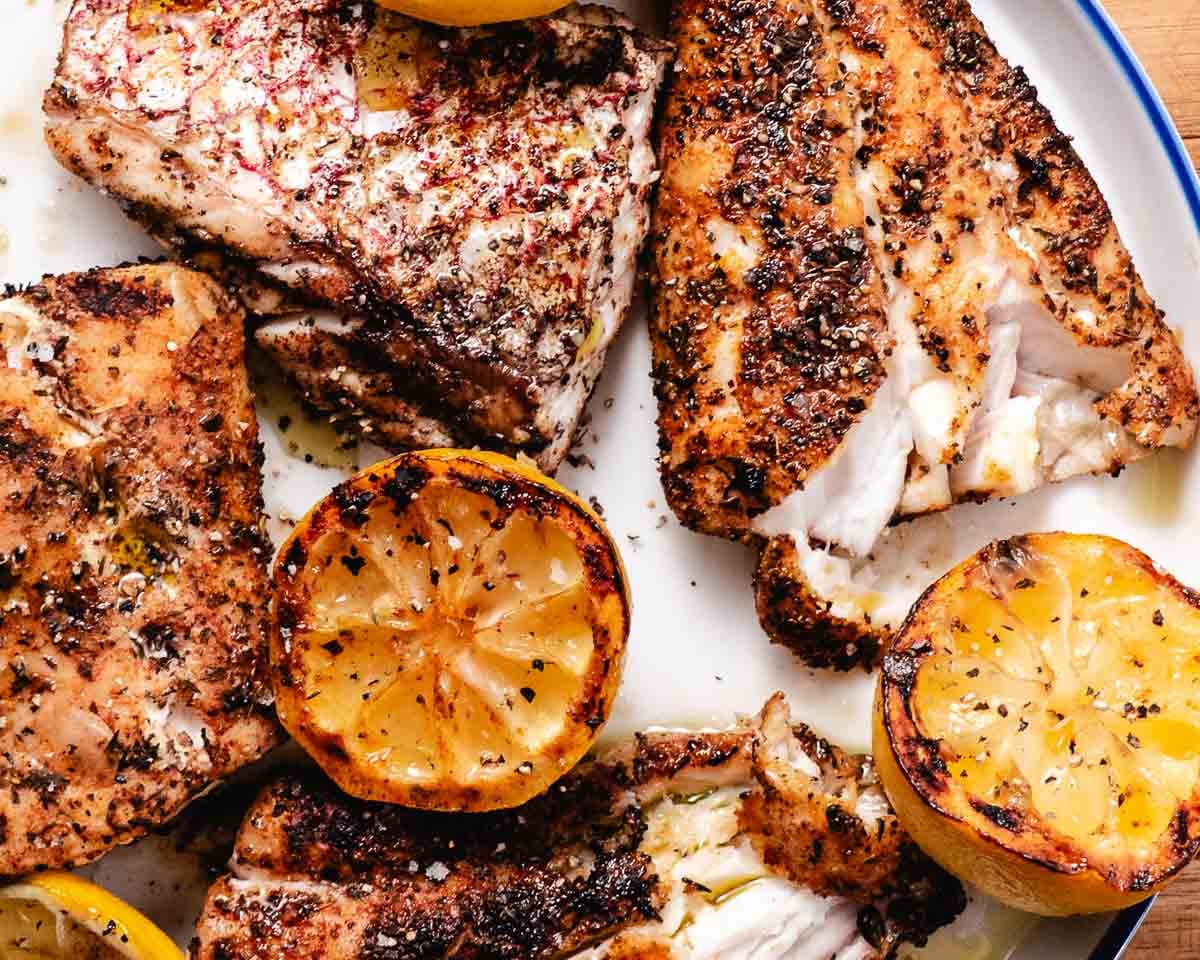 Grilled red snapper fillets with charred lemons and blackened seasoning on a white platter.