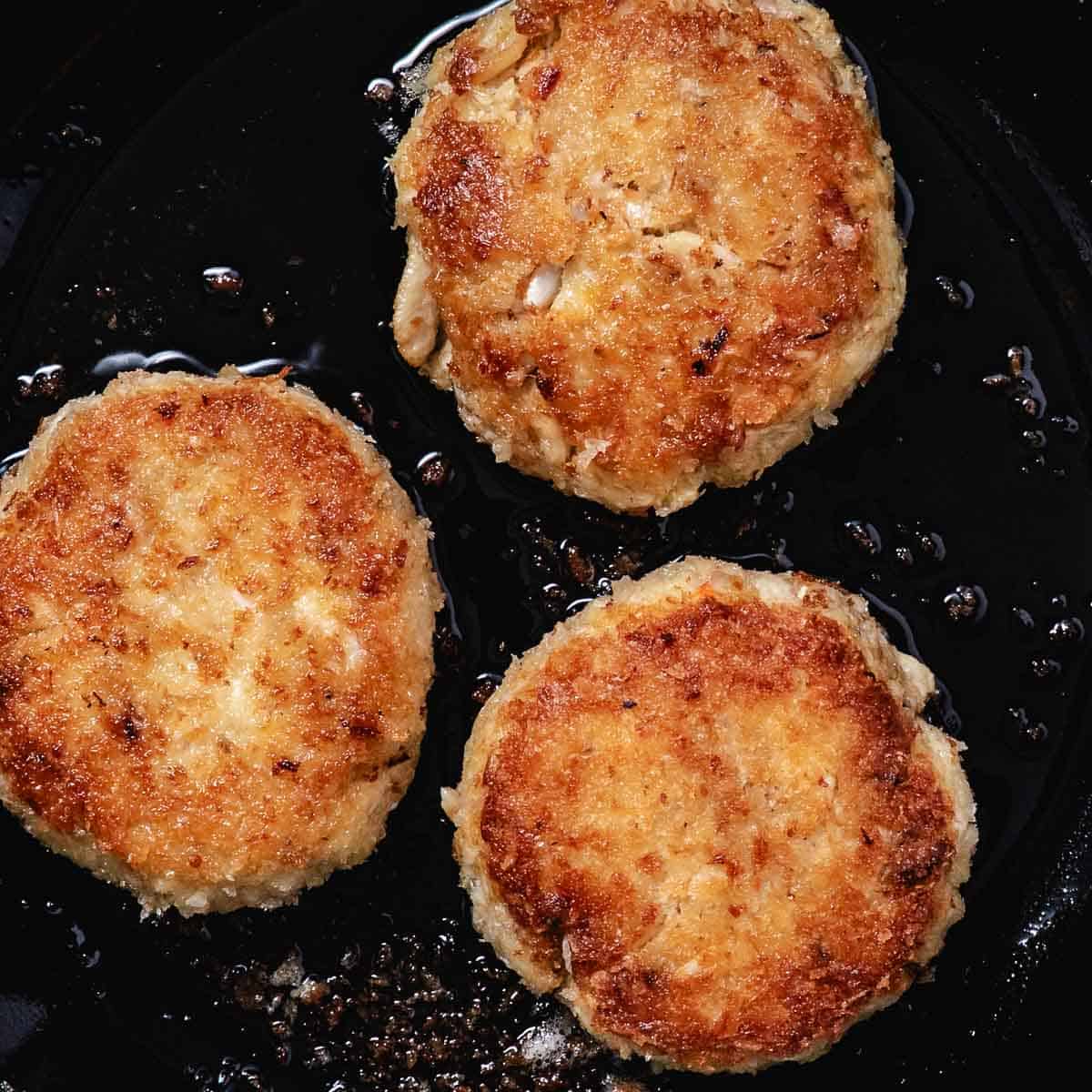 Three panko crab cakes pan-frying until crisp and golden brown.