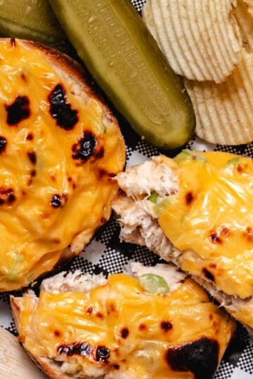 Open-faced tuna melt on a bagel with pickles and chips on deli paper.