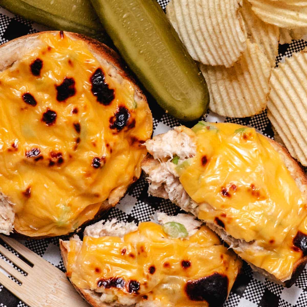 Open-faced tuna melt on a bagel with pickles and chips on deli paper.