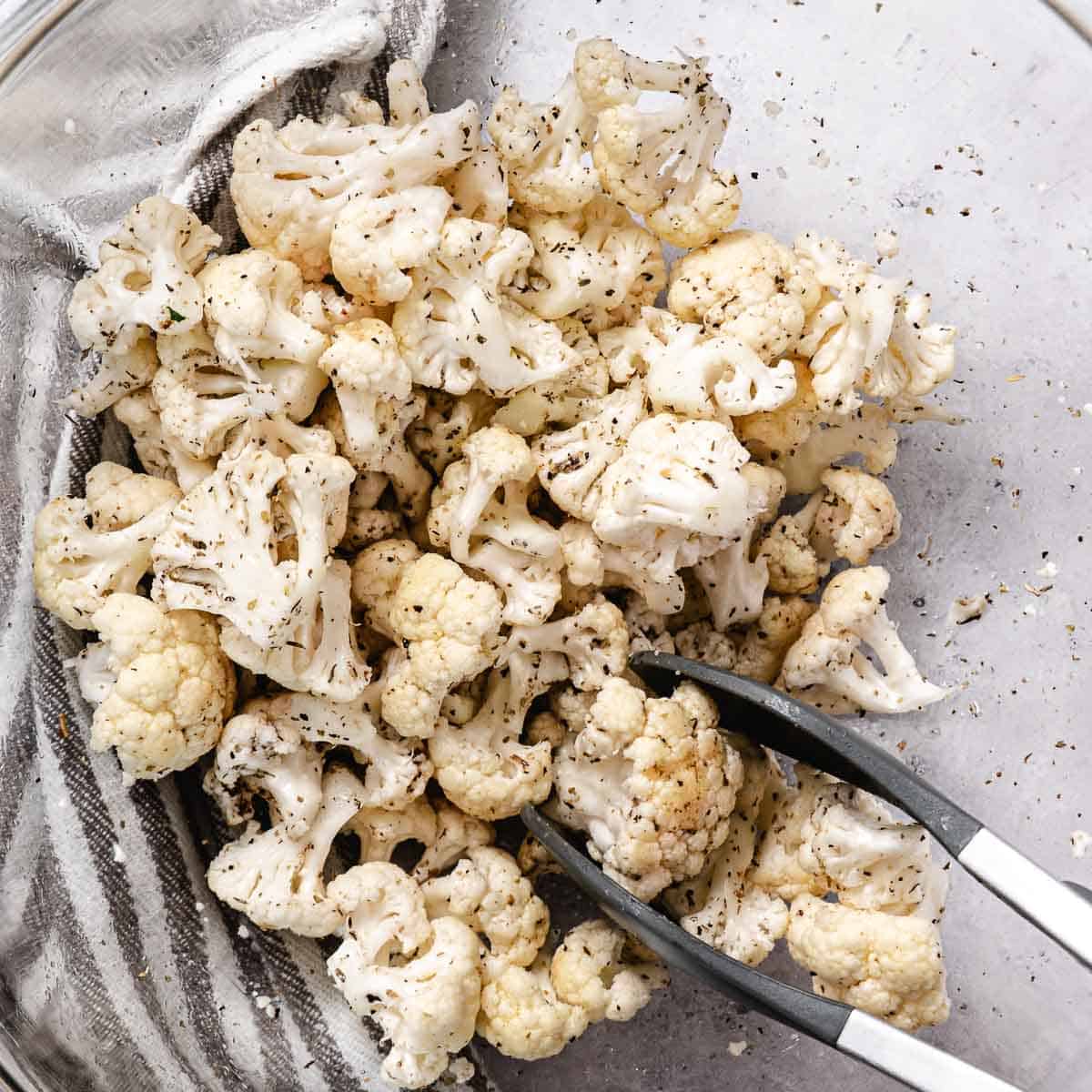 Cauliflower florets tossed with olive oil, garlic, Italian seasoning, salt, and pepper in a glass bowl.