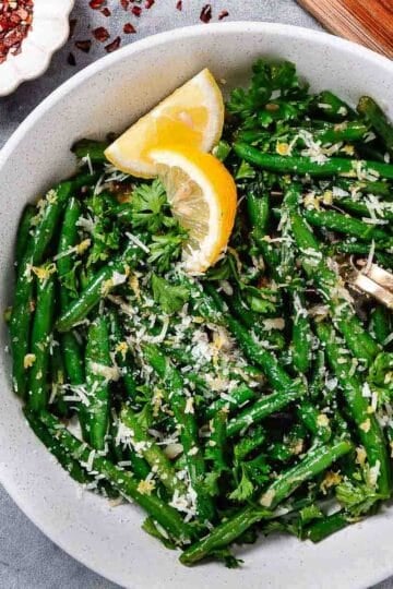 overhead view of green beans with lemon zest and parmesan cheese. Lemon wedges and chili flakes to the side.