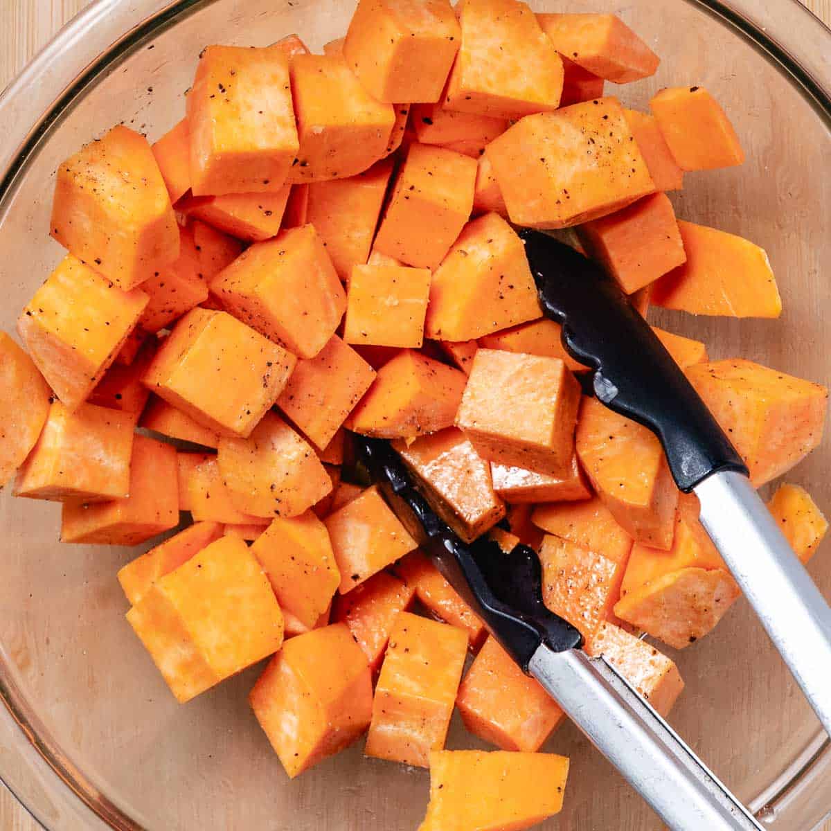 Mixing baked sweet potato cubes with oil and seasonings with tongs.