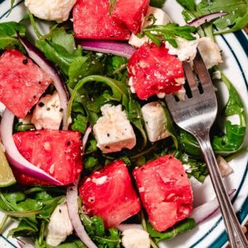 Watermelon arugula salad with feta and red onion on a platter, topped with lime vinaigrette, with a fork lifting a watermelon cube from the salad.