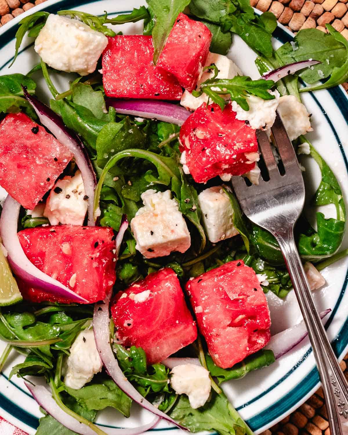 Watermelon arugula salad with feta and red onion on a platter, topped with lime vinaigrette, with a fork lifting a watermelon cube from the salad.