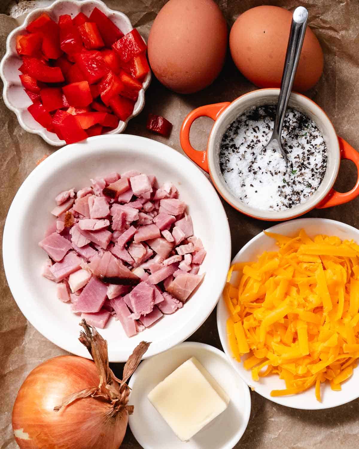 Overhead view of Denver omelet ingredients laid out, including eggs, ham, peppers, onions, cheese, butter, salt, and pepper.