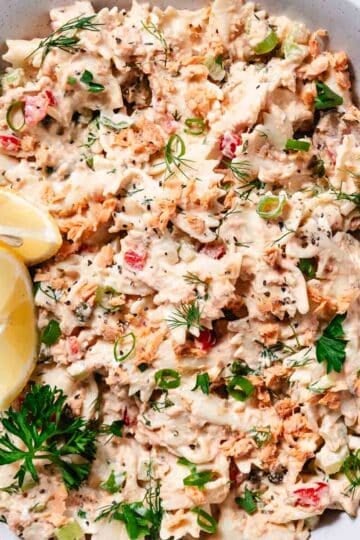 Overhead shot of salmon pasta salad made with bowtie pasta, salmon, capers, celery, fresh herbs, and a creamy dressing, served with lemon.
