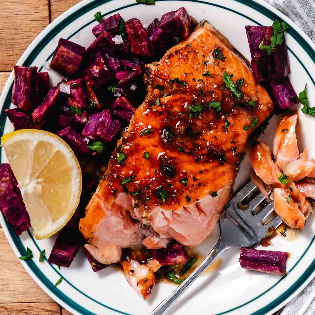 Honey garlic glazed salmon on a plate with roasted purple sweet potatoes and a lemon wedge. A fork is pulling off a few flakes of salmon.