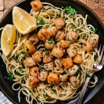 saut&eacute;ed bay scallops served over spaghetti with parsley and lemon wedges on a black plate.