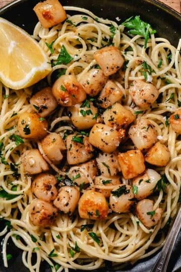 saut&eacute;ed bay scallops served over spaghetti with parsley and lemon wedges on a black plate.