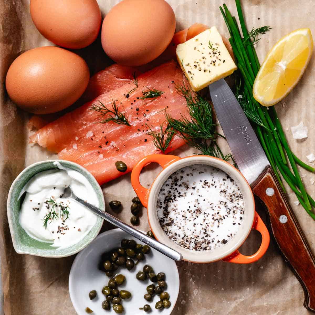 Overhead view of smoked salmon omelet ingredients on parchment paper, including whole eggs, sliced smoked salmon with dill, sour cream, capers, butter on a knife, chives, lemon wedge, and a bowl of salt and pepper.