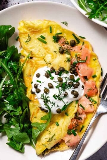 Smoked salmon omelet folded and plated on a white dish, finished with a dollop of sour cream, capers, dill, and chives, with fresh arugula on the side and a fork alongside.