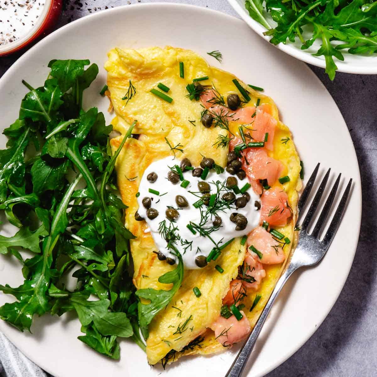 Smoked salmon omelet folded and plated on a white dish, finished with a dollop of sour cream, capers, dill, and chives, with fresh arugula on the side and a fork alongside.