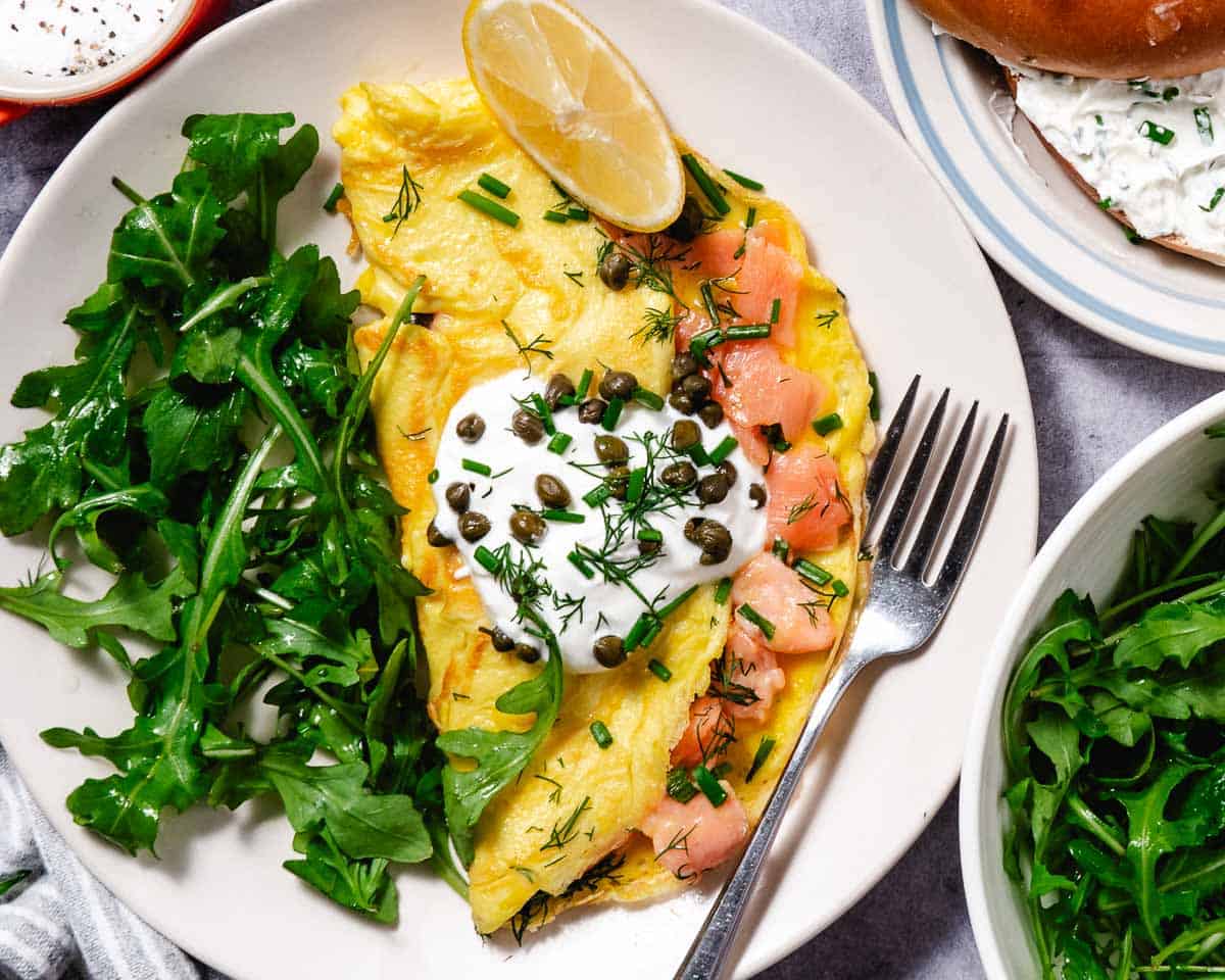 Overhead view of a smoked salmon omelet topped with sour cream, capers, dill, and chives, served with a lemon wedge and a side of fresh arugula on a white plate with a fork.