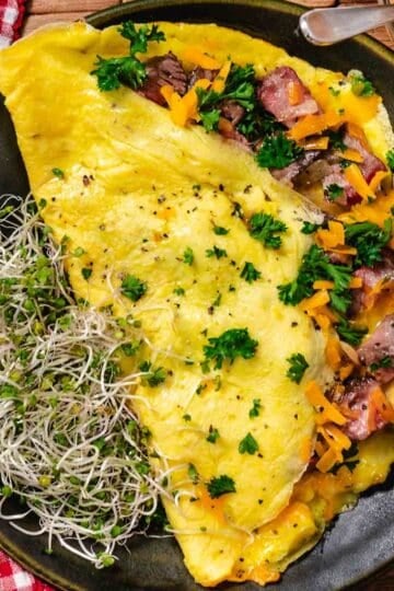 High-protein steak omelet cooked in butter with cheddar cheese and onions, folded and plated with raw broccoli sprouts for a simple breakfast.