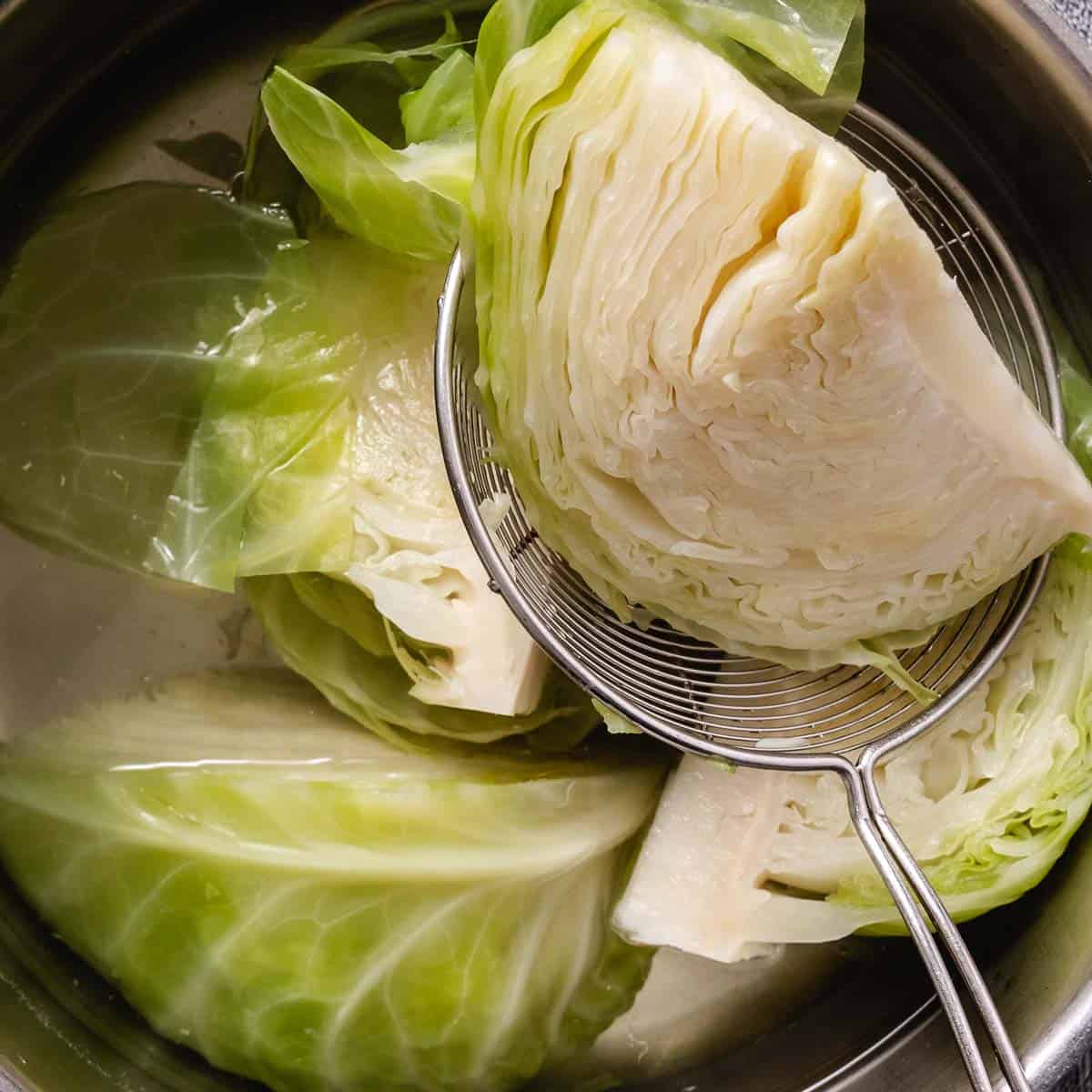 Boiled cabbage wedges draining in a pot of hot water with a metal strainer.