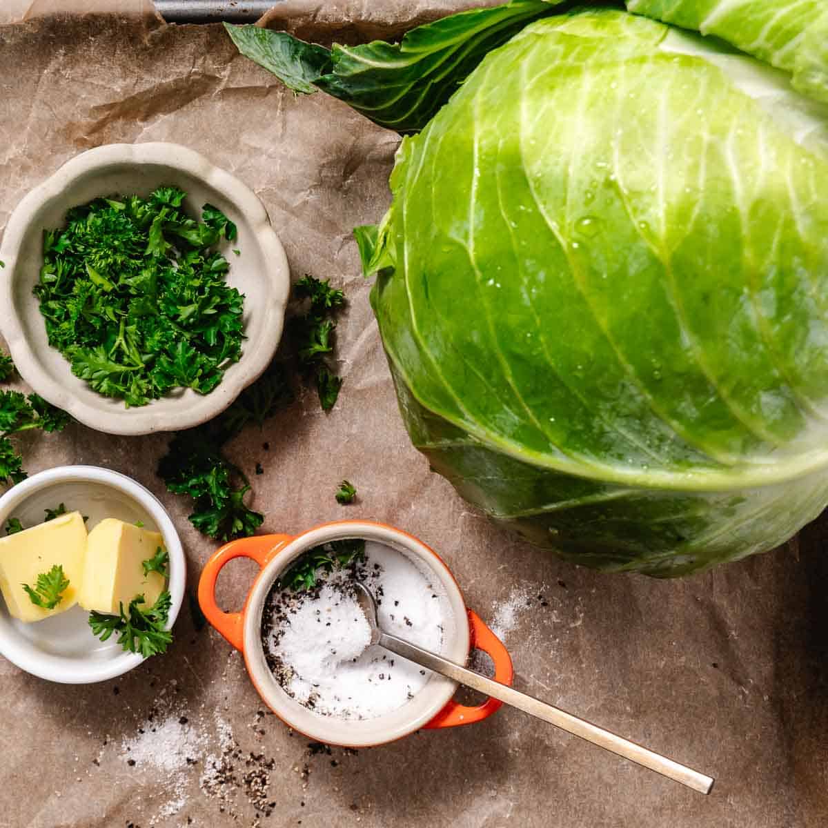 Whole green cabbage with butter, salt, pepper, and chopped parsley on parchment paper.