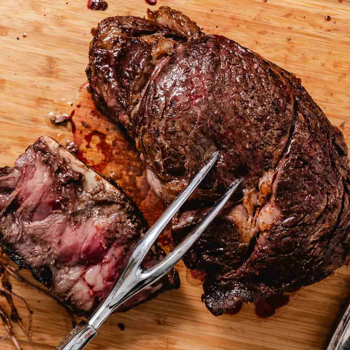 Small standing rib roast resting on a cutting board, browned and seared in a cast iron skillet, with one slice cut to show a pink, tender center.